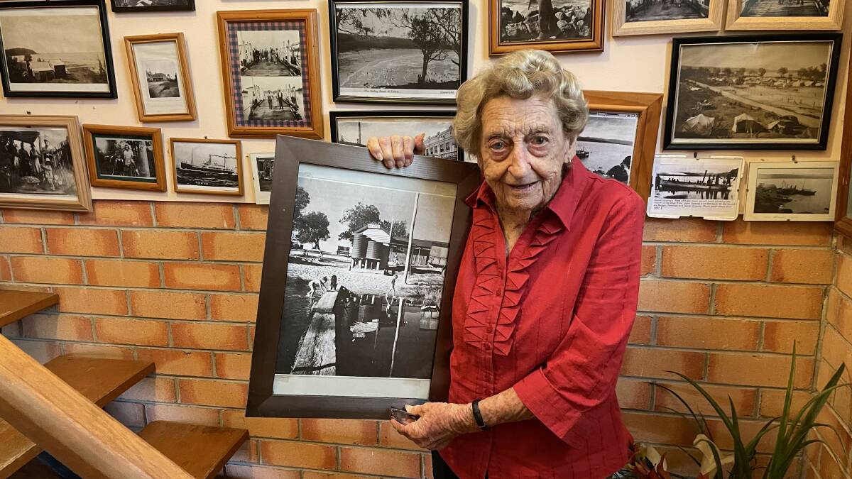 Betty Koellner smiling with an image of Tathra Surf Lifesaving club taken in the 1940s. Picture by James Parker